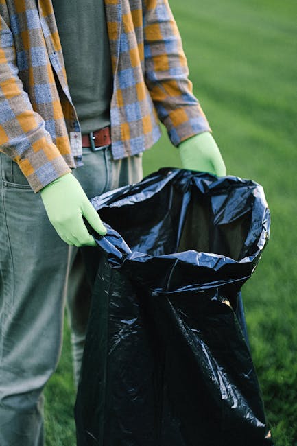 A waste collection worker wearing a blue uniform with an orange safety vest and a matching blue cap is seen standing on a cobbled street, emptying a blue wheeled bin into the rear opening of a white r